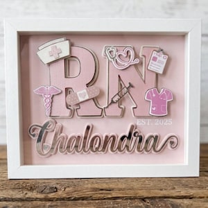 May include: A framed pink and white RN shadow box with medical-themed embellishments. The design includes a nurse's cap, stethoscope, and syringe. The name "Chalondra" is displayed below the letters, with "EST. 2025" at the bottom.