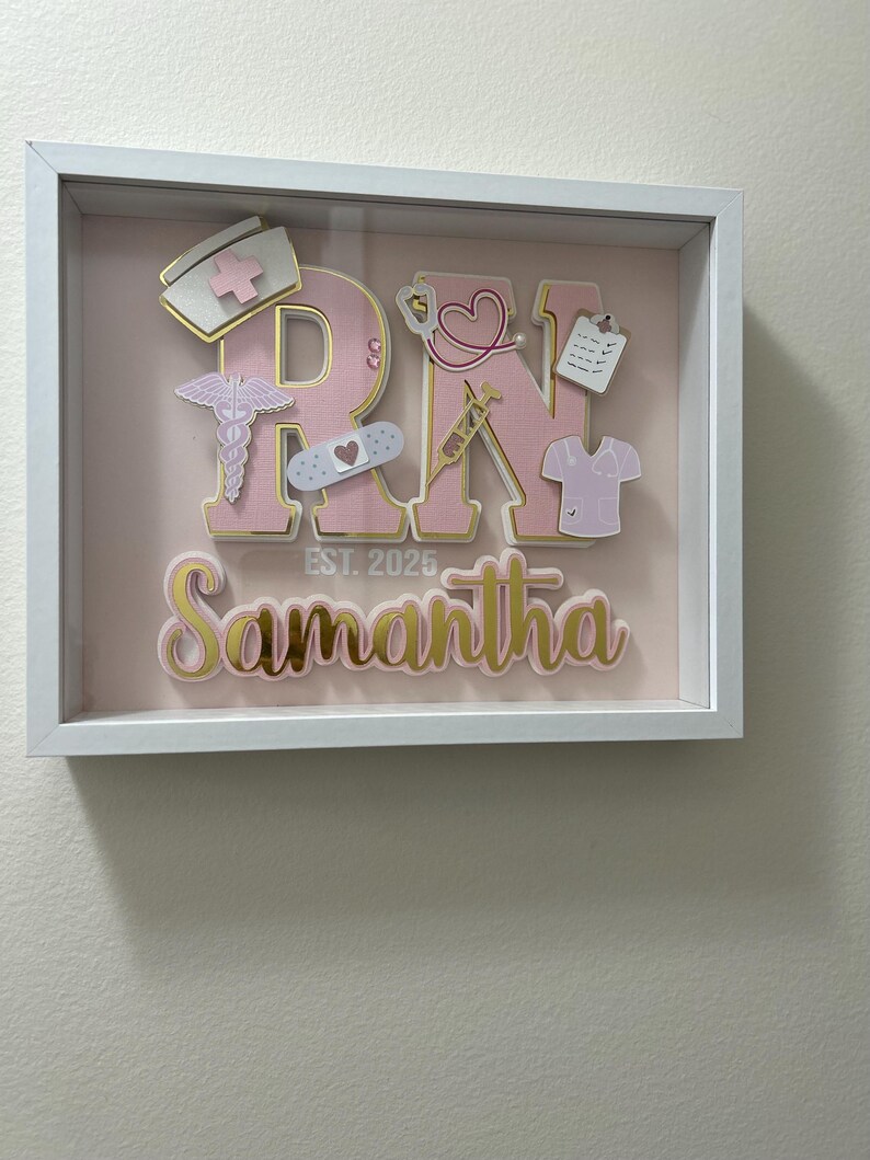 May include: A white shadow box with a pink background and the letters "RN" in pink with gold trim. The letters are decorated with various medical-themed cutouts, including a stethoscope, a bandage, a syringe, and a heart. The name "Samantha" is written below the letters in pink with gold trim. The text "EST. 2025" is written below the letters.
