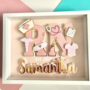 May include: A pink and gold framed shadow box with the letters RN in pink with gold trim. The letters are decorated with various medical symbols and a pink scrub top. The name Samantha is written below the letters with the text EST. 2025.