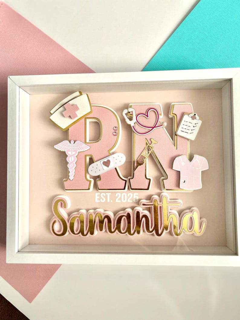 May include: A pink and gold framed shadow box with the letters RN in pink with gold trim. The letters are decorated with various medical symbols and a pink scrub top. The name Samantha is written below the letters with the text EST. 2025.