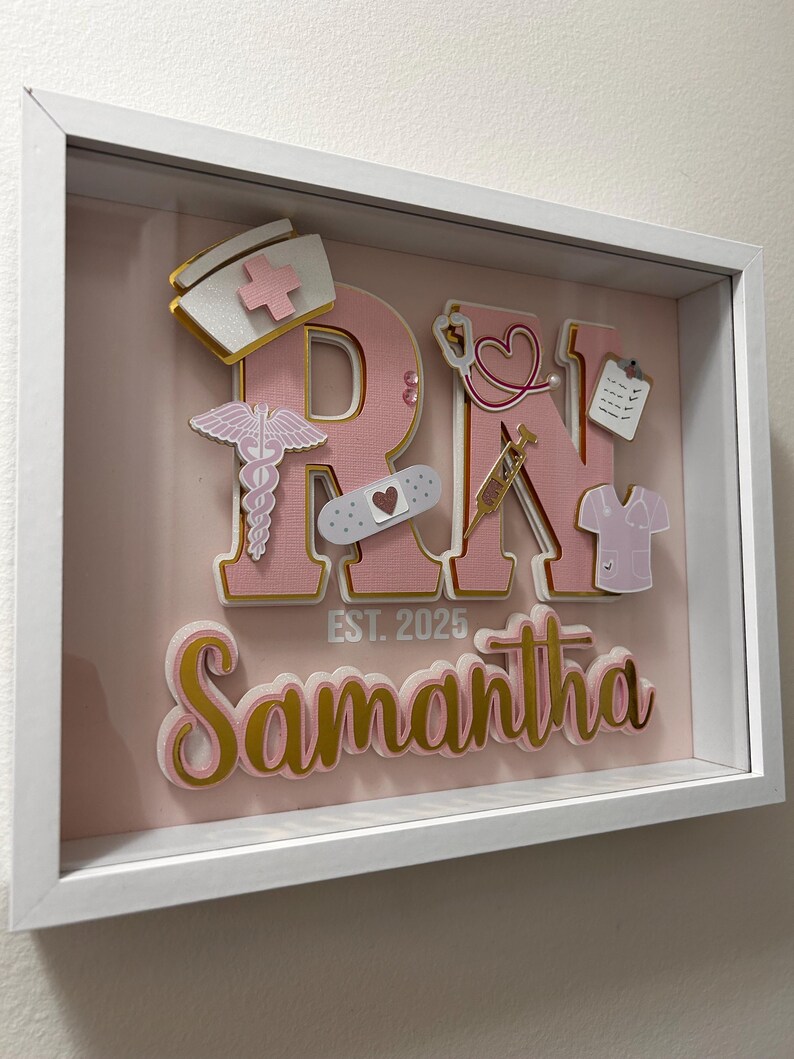 May include: A pink and gold framed shadow box with the letters "RN" in pink with gold trim. The letters are decorated with various medical symbols and a white bandage. The name "Samantha" is written below the letters with the text "EST. 2025" above the name.