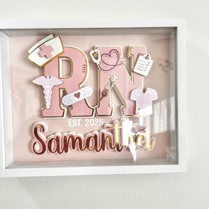 May include: A pink and gold shadow box with the letters "RN" in large pink letters with gold trim. The letters are decorated with small pink and gold medical symbols. The name "Samantha" is written in gold letters below the letters "RN". The text "EST. 2025" is written in gold letters below the name.
