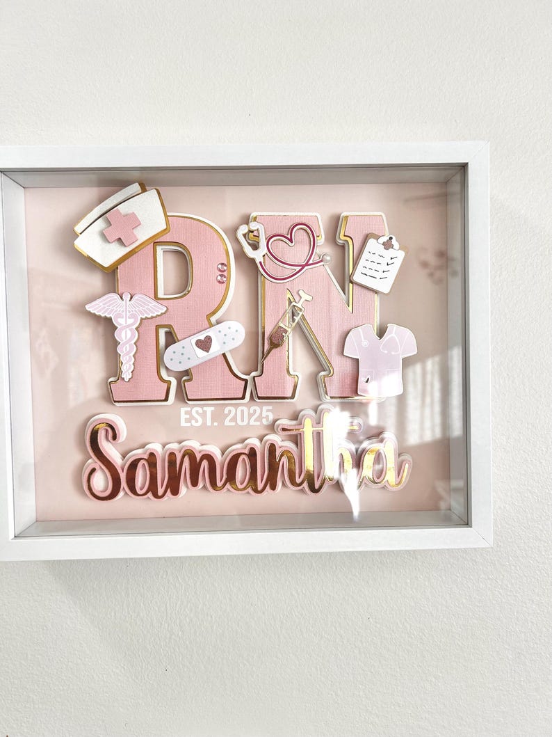 May include: A pink and gold shadow box with the letters "RN" in large pink letters with gold trim. The letters are decorated with small pink and gold medical symbols. The name "Samantha" is written in gold letters below the letters "RN". The text "EST. 2025" is written in gold letters below the name.