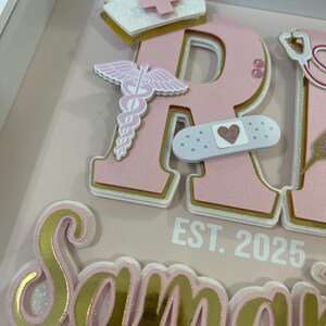 May include: Pink and gold glitter letters spelling "RN" with a white nurse's hat, a pink caduceus, a pink and white bandage with a pink heart, and the text "EST. 2025" below. The letters are layered and have a white outline.
