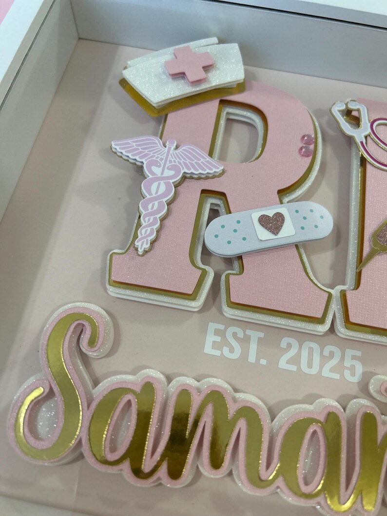 May include: Pink and gold glitter letters spelling "RN" with a white nurse's hat, a pink caduceus, a pink and white bandage with a pink heart, and the text "EST. 2025" below. The letters are layered and have a white outline.
