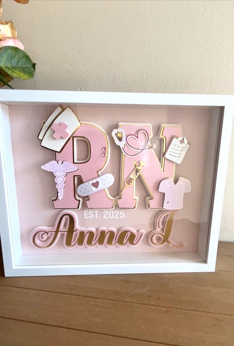 May include: A pink and gold framed shadow box with the letters "RN" in pink with gold trim. The letters are decorated with various medical symbols and tools, including a stethoscope, a bandage, a syringe, and a nurse's cap. The text "EST. 2025" is below the letters. The name "Anna L" is written in pink and gold lettering below the date.