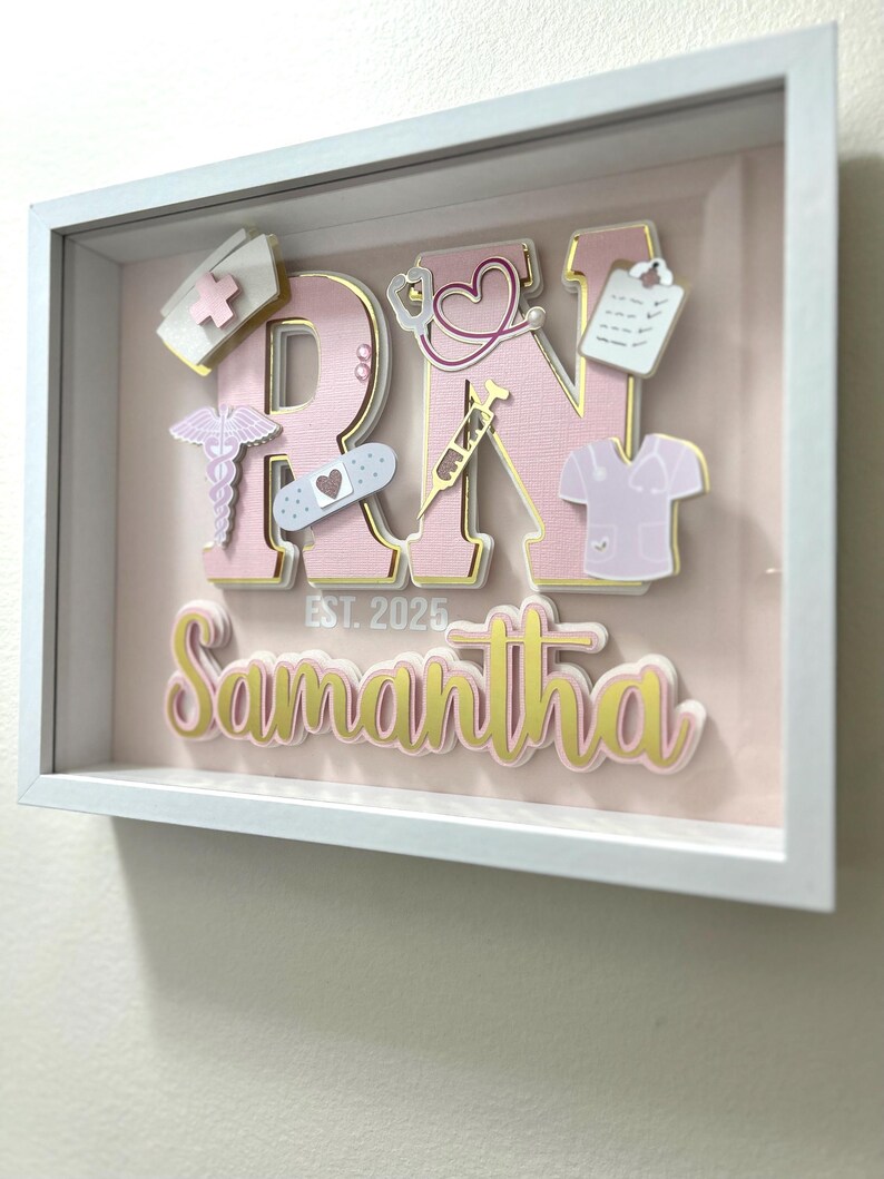May include: A pink and gold framed shadow box with the letters "RN" in pink with gold trim. The letters are decorated with various medical symbols and the name "Samantha" is written below the letters with the text "EST. 2025".