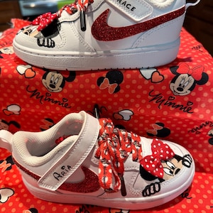 May include: White and red toddler sneakers with a Minnie Mouse design. The sneakers feature red glitter accents, Minnie Mouse graphics, and the names "Grace" and "Aria" on the straps. The laces are red with white polka dots.