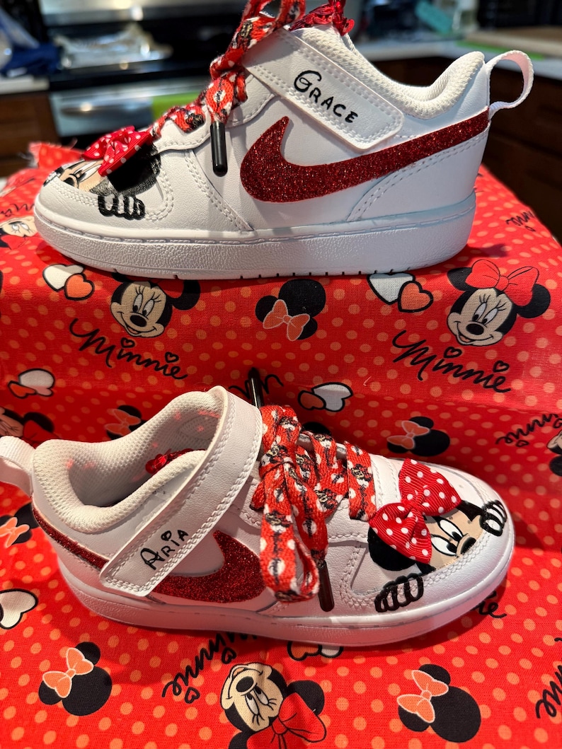 May include: White and red toddler sneakers with a Minnie Mouse design. The sneakers feature red glitter accents, Minnie Mouse graphics, and the names "Grace" and "Aria" on the straps. The laces are red with white polka dots.