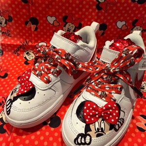 May include: White sneakers with red accents and Minnie Mouse graphics. The shoes feature red and white polka dot bows and laces, with a matching red background. The shoes have a velcro strap and are decorated with sequins.