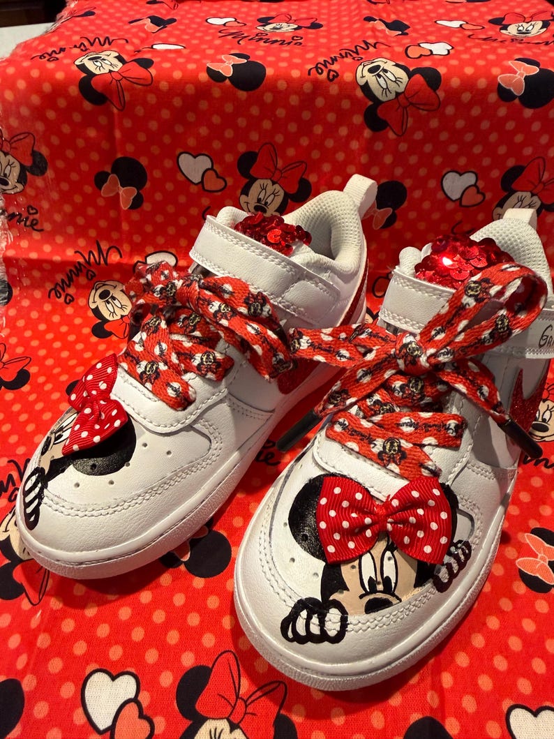 May include: White sneakers with red accents and Minnie Mouse graphics. The shoes feature red and white polka dot bows and laces, with a matching red background. The shoes have a velcro strap and are decorated with sequins.