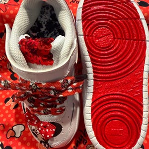 May include: White and red toddler sneakers with Minnie Mouse accents. The sneakers feature red laces with Minnie Mouse silhouettes, a red bow, and a red sole. The interior is white with a black and white pattern.