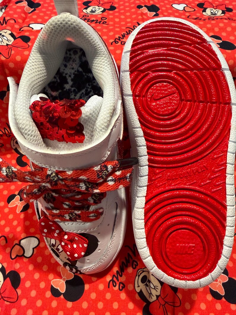 May include: White and red toddler sneakers with Minnie Mouse accents. The sneakers feature red laces with Minnie Mouse silhouettes, a red bow, and a red sole. The interior is white with a black and white pattern.