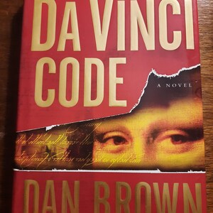 May include: A red hardcover book with the title "The Da Vinci Code" in gold lettering. The book is by Dan Brown and is a novel. The cover features a close-up of the Mona Lisa's eyes.