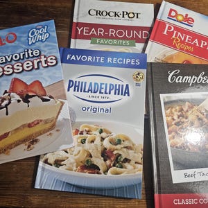 May include: A collection of five hardcover cookbooks featuring branded titles including Jell-O Favorite Desserts, Crock-Pot Year-Round Favorites, Dole Pineapple Recipes, Philadelphia Original Favorite Recipes, and Campbell's Beef Taco Skillet Classic Comfort Food.