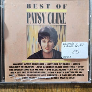 May include: A CD case featuring the album "Best of Patsy Cline." The cover displays a portrait of Patsy Cline with the album title in bold letters. The tracklist includes "Walkin' After Midnight" and "I Can See an Angel."