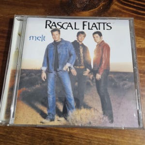 May include: A Rascal Flatts Melt music CD in a clear plastic jewel case. The cover features three individuals standing in a desert landscape at sunset. The text Rascal Flatts is printed in black at the top, with the word melt in blue on the left.