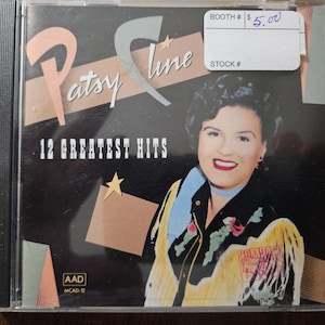 May include: A compact disc case featuring Patsy Cline's "12 Greatest Hits." The album cover displays a portrait of Patsy Cline with a black background, the title in white, and a star graphic. A sticker with a price tag is on the top right.
