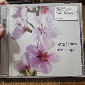 May include: A CD case featuring a floral design with light pink blossoms. The text "etta james love songs" is visible. The CD is in a clear plastic case, with a price tag in the upper right corner.
