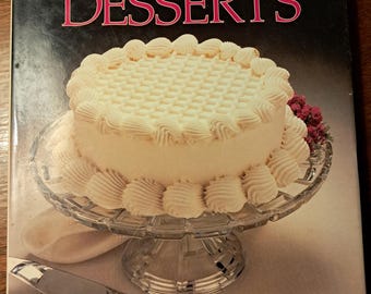 May include: A cookbook titled "Elegant Desserts" with a white cake with white frosting on a glass cake stand. The cake has a grid pattern on the top and swirls of frosting around the bottom.