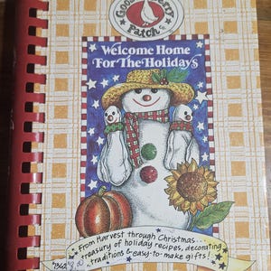 May include: A spiral-bound cookbook titled Welcome Home For The Holidays by Gooseberry Patch Co. The cover features a snowman wearing a straw hat and plaid scarf, a pumpkin, and a sunflower. Text reads From Harvest through Christmas.