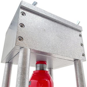 May include: A silver metal press with a red cylinder and a textured surface. The press has four legs and is designed for pressing objects.