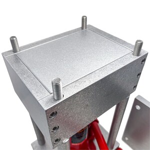 May include: A silver metal press with a rectangular top plate and four bolts. The press is designed for pressing items such as jewelry or metal.