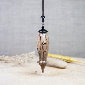 May include: A wooden pendulum with a black top and a pointed bottom, suspended by a dark cord. The pendulum has a light brown and black wood grain pattern. The background is a neutral color, with dried plants on a wooden surface.