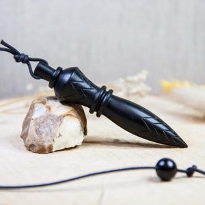 Könnte beinhalten: Ein schwarzes Holzpendel mit spitzem Ende und dekorativen Schnitzereien. Das Pendel ist an einer schwarzen Schnur aufgehängt und liegt auf einem Stein. Der Hintergrund ist hell und neutral.