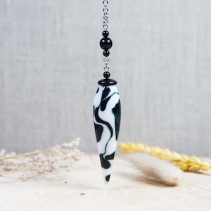 Peut inclure: Un pendule avec un motif marbré noir et blanc. Le pendule allongé en forme de goutte d'eau est suspendu à une chaîne argentée avec des perles noires. L'arrière-plan est de couleur gris clair neutre.