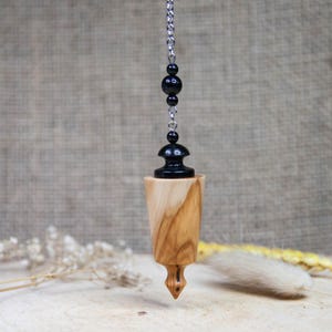 May include: A wooden pendulum with a black metal top and a chain with black beads. The pendulum is on a light brown wooden surface.