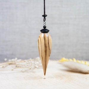 May include: A wooden pendulum with a teardrop shape, featuring light brown and dark brown wood grain. The pendulum is suspended from a black cord with a small black bead and a black cap. The background is a neutral gray.
