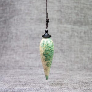 May include: A teardrop-shaped pendant with a green and cream marbled pattern. The pendant is suspended from a black cord with a knot and a black cap. The background is a neutral gray.