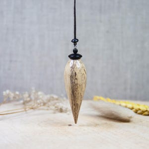 May include: A wooden pendulum with a light brown and gray wood grain pattern. The pendulum is suspended from a black cord and has a black metal top.
