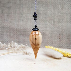 May include: A wooden pendulum with a pointed end, suspended by a silver chain and black beads. The pendulum is light brown with a natural wood grain pattern. The background is a neutral, textured fabric.