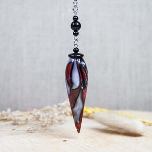 May include: A pendulum with a teardrop-shaped body, featuring a blend of red, white, and black colors. It hangs from a silver chain with black beads. The background is a neutral gray, with dried plants on a wooden surface.