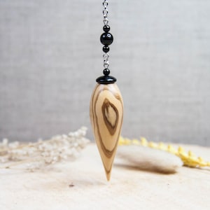 May include: A pendulum with a teardrop-shaped wooden body, featuring a light brown and dark brown wood grain pattern. The pendulum is suspended by a silver chain with black beads and a black cap.