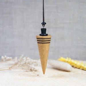 Puede incluir: Un péndulo de madera con una punta puntiaguda y detalles negros. El péndulo está colgado de un cordón negro.