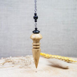 May include: A wooden pendulum with a silver chain and black beads. The pendulum has a light brown, conical shape with a rounded top and a pointed bottom. The background is a neutral beige.