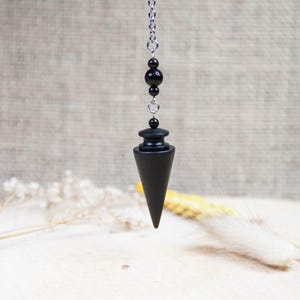 Peut inclure: Pendule noire avec une chaîne argentée et des perles noires. Le pendule est en forme de cône avec un sommet arrondi.