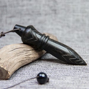 Puede incluir: Colgante de madera negra con un extremo puntiagudo y un diseño de chevrón tallado. El colgante está suspendido por un cordón marrón oscuro y descansa sobre un trozo de madera flotante. Una pequeña cuenta negra también está unida al cordón.