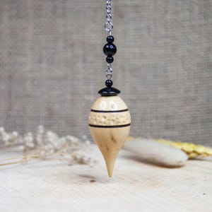 May include: A wooden pendulum with a light brown color and black accents. The pendulum is carved with a decorative pattern and hangs from a silver chain with black beads.
