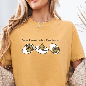 May include: Mustard yellow t-shirt with the phrase "You know why I'm here." and a graphic of three deviled eggs. The eggs have a brown filling and are on a white background. The shirt is a casual style.