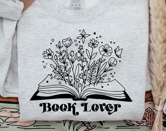 Puede incluir: Una sudadera gris con una ilustración en blanco y negro de un libro abierto con flores que salen de él. El texto "Book Lover" está impreso debajo del libro.