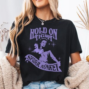May include: Black t-shirt with a purple graphic design that says "Hold On Tight Spider Monkey" with a cartoon image of a woman holding another woman's head.