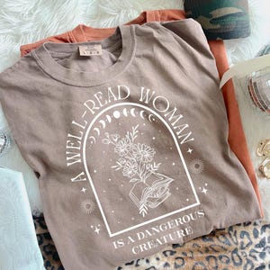 May include: A taupe-colored t-shirt with the text "A WELL-READ WOMAN IS A DANGEROUS CREATURE" in white. The graphic includes an open book, flowers, and celestial elements. Additional orange t-shirt and a camouflage hat are also visible.