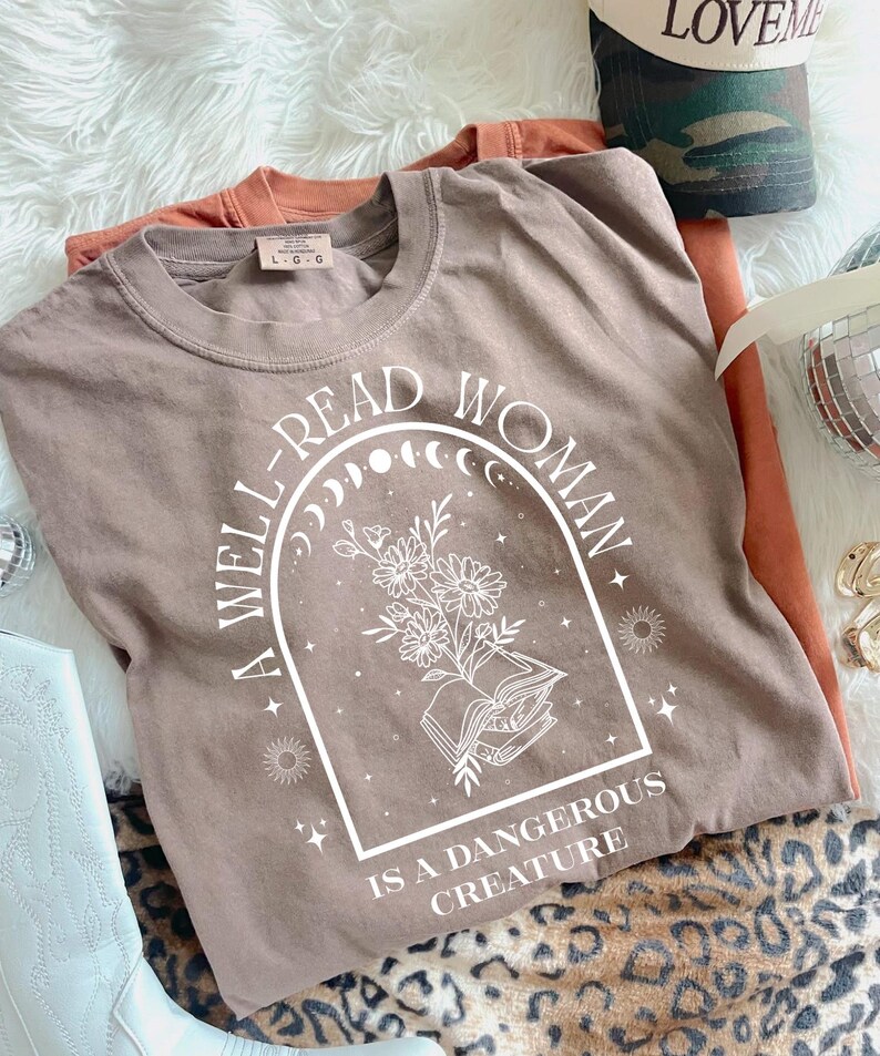 May include: A taupe-colored t-shirt with the text "A WELL-READ WOMAN IS A DANGEROUS CREATURE" in white. The graphic includes an open book, flowers, and celestial elements. Additional orange t-shirt and a camouflage hat are also visible.