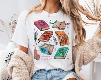 May include: White t-shirt featuring a graphic print of colourful books, feathers, candles, and bats. The text on the shirt reads "Don't just read for one moment about doing what brings you joy."