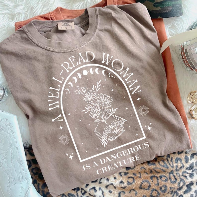 May include: A brown t-shirt with the text "A WELL-READ WOMAN IS A DANGEROUS CREATURE" in a white arched design. The design includes flowers, a book, and celestial elements. Another orange t-shirt is visible in the background.