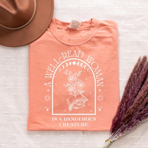 May include: Coral-colored t-shirt with the text "A WELL-READ WOMAN IS A DANGEROUS CREATURE" in white. The design includes an arched frame with a floral and book illustration. A brown hat and a bundle of dried flowers are also visible.